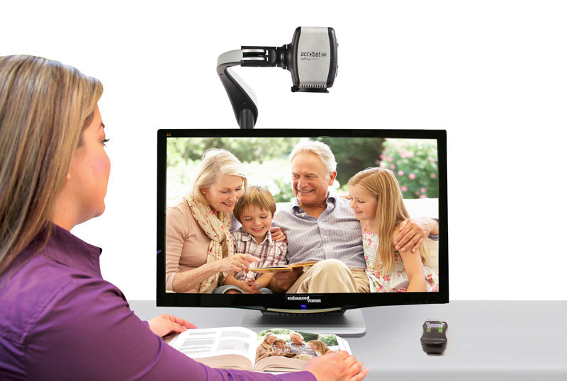 A woman sits in front of the Acrobat HD Ultra 27" screen. The 3-1 camera attached to a metal arm above the screen is margnifing the image of a family of four reading a book. A black remote with white and green button sits to the right of the open book