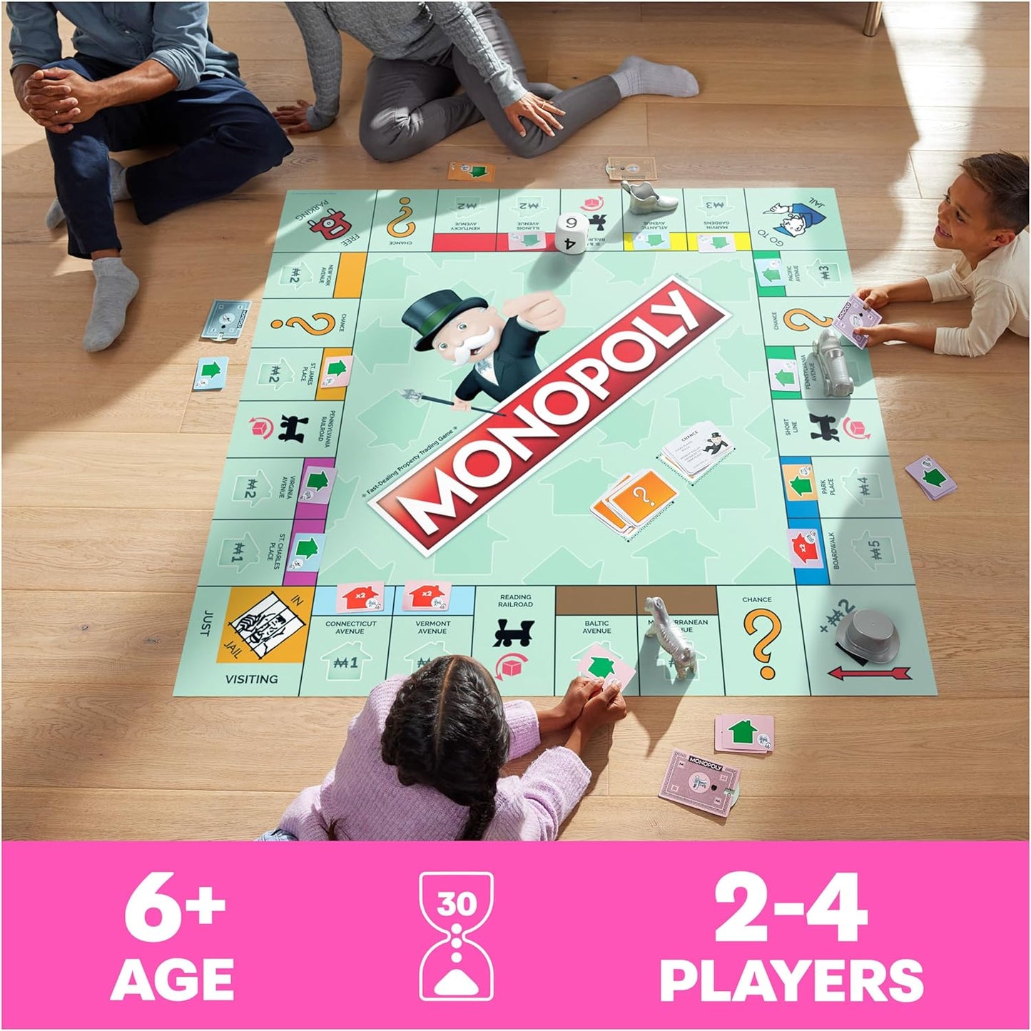 Four kids sitting around the Giant Monopoly Board game holding large cards and paper money inside their house. You can see the sun shining in the window and reflecting on the ground where they are playing.