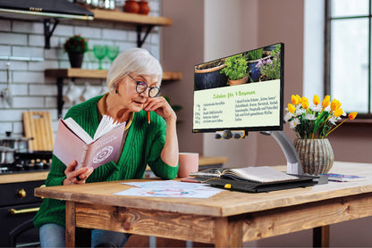 A woman is sitting at a table in a kitchen looking over a book and viewing a recipe that is magnified in true color on the Mezzo 24 in electronic magnifier that is sitting off to the right.