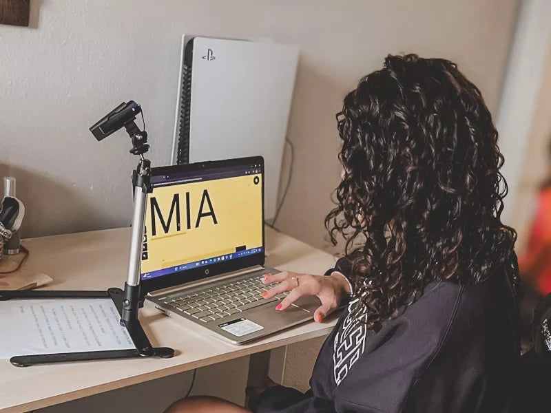 A woman using the OrCam Magnifier and Text Scanner feature on her laptop with the device placed into a tall stand pointing down at text on the table.