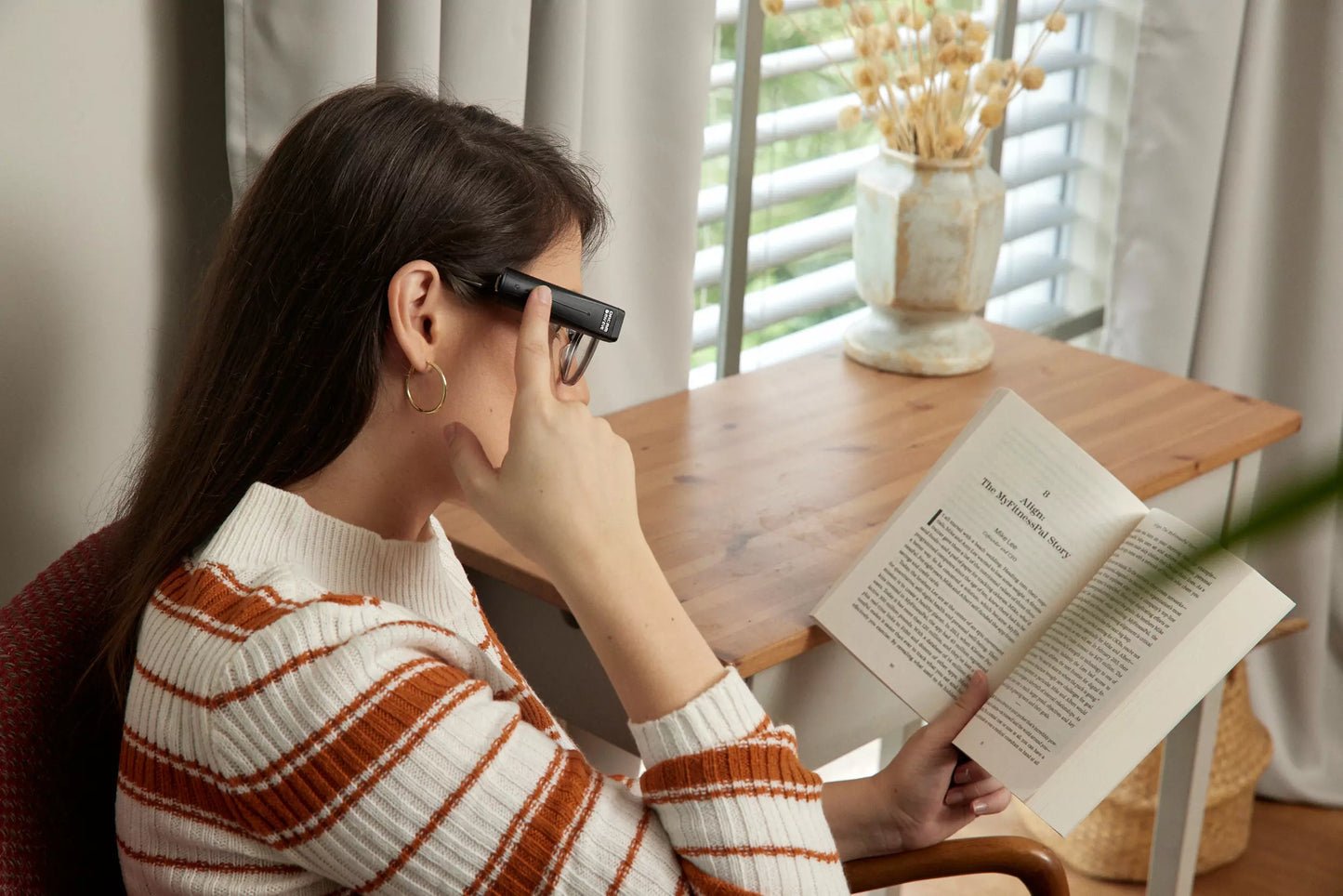 A woman wearing the OrCam on her glasses and holding a book while tapping the side of the device.